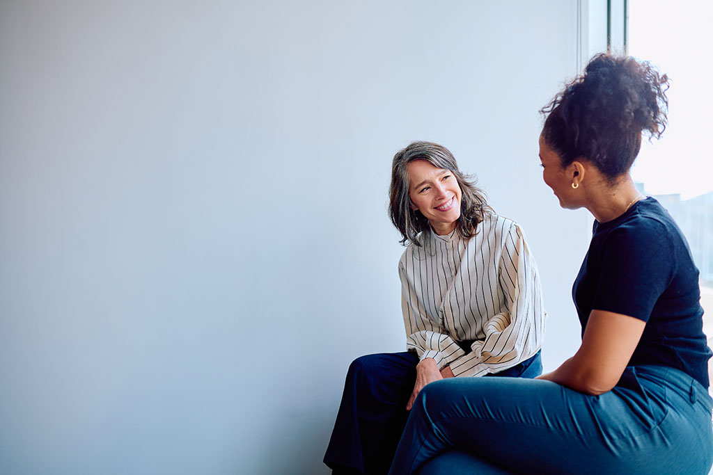 Two diverse businesswomen smiling during an informal meeting, engaging in a pleasant conversation about work in a bright workplace conflict coaching